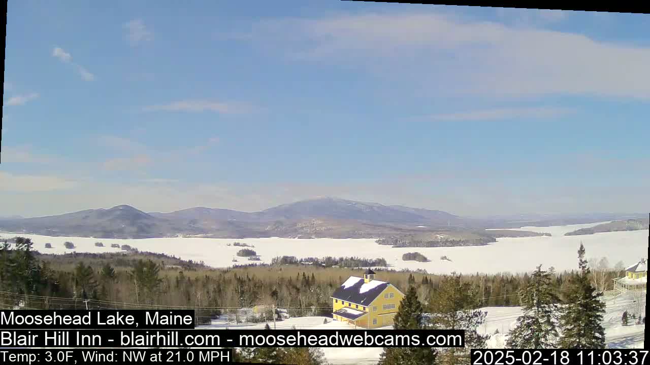 A snow-covered landscape with a yellow building, a frozen lake, and distant mountains under a clear blue sky.