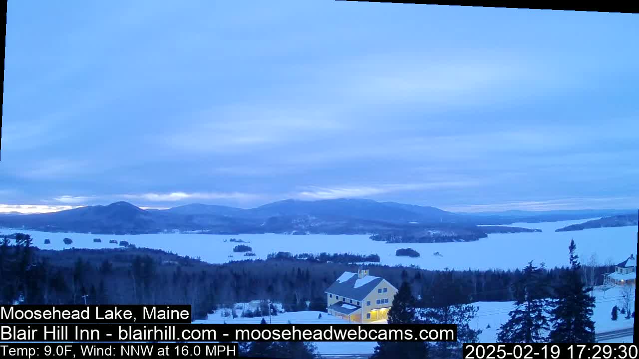 A snow-covered lake and distant mountains are visible under a pale blue, partly cloudy sky, with a building situated in a snowy wooded area in the foreground.