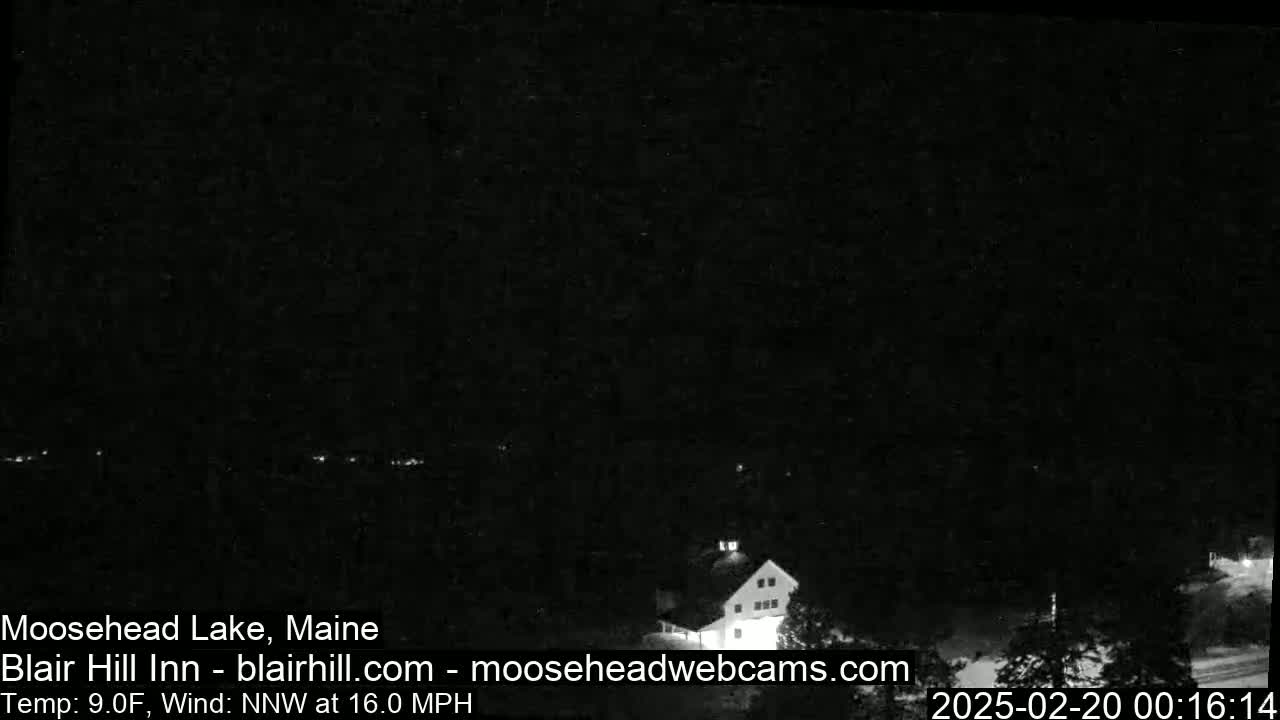 A nighttime, low-light view of a building near a dark, tree-lined lake under a dark sky with a northwest wind at 16 mph and a temperature of 9°F.