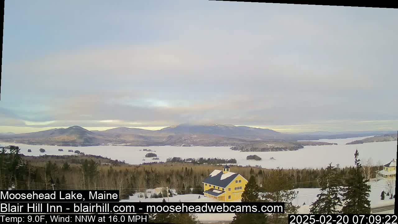 A partly cloudy, cold day overlooks a snow-covered lake and distant mountains, with a yellow building nestled in the foreground amongst evergreens.