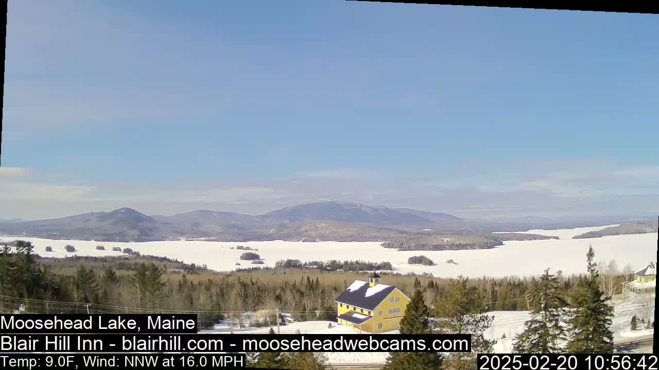 A yellow building sits on a snow-covered hill overlooking a frozen lake and distant mountains under a clear blue sky.