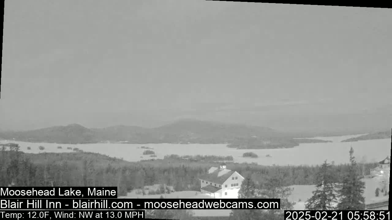 A snow-covered landscape overlooks a large, partially frozen lake under a gray sky, with a building visible in the foreground.