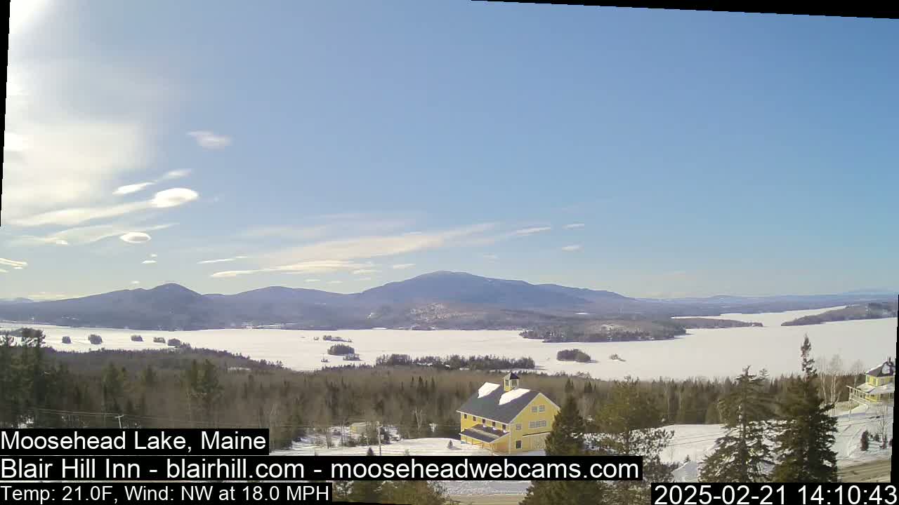 A partly sunny, cold day with a northwest wind shows a frozen lake surrounded by snow-covered trees and mountains, with a yellow building in the foreground.