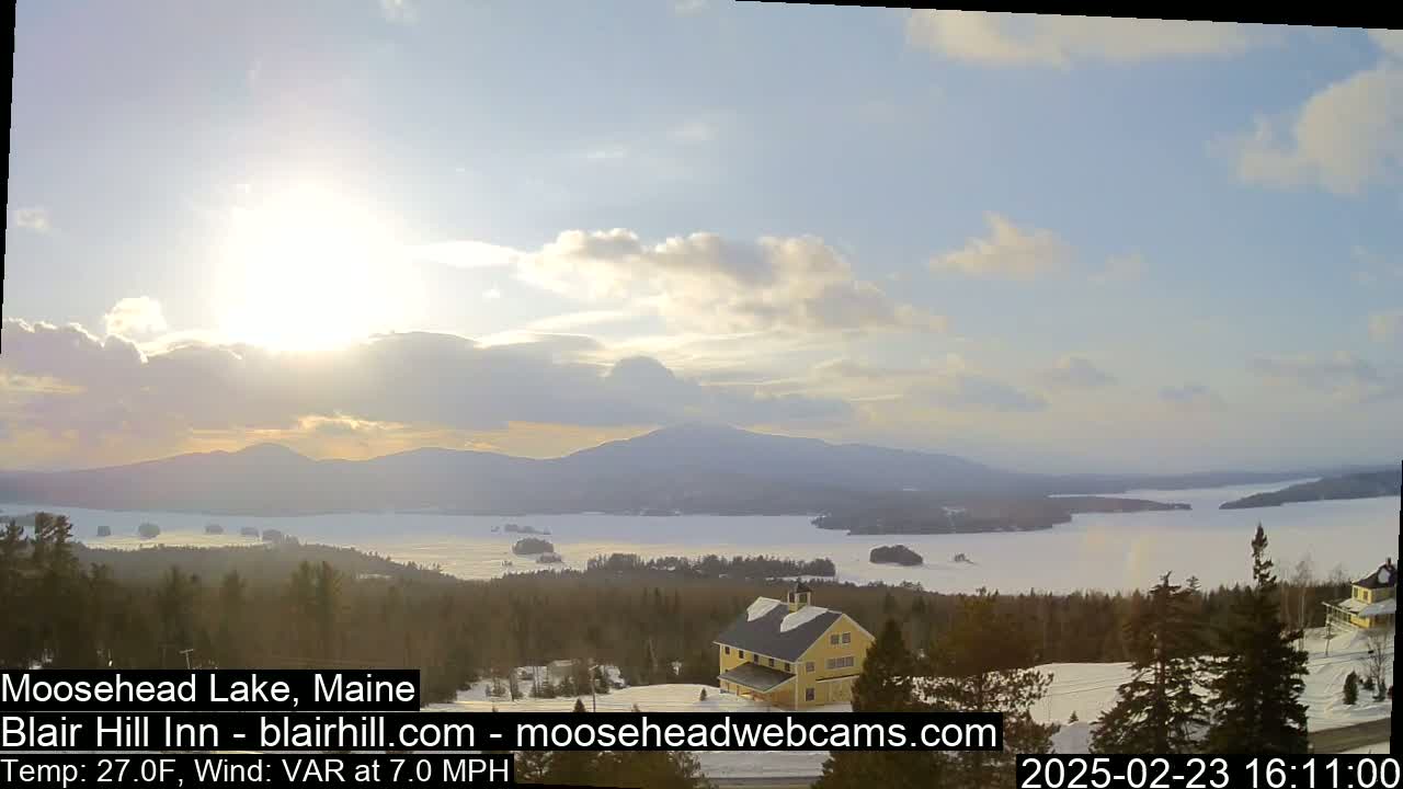 A partly cloudy, sunny day overlooks a snow-covered lake and distant mountains, with a yellow building situated on a snow-covered hillside in the foreground.