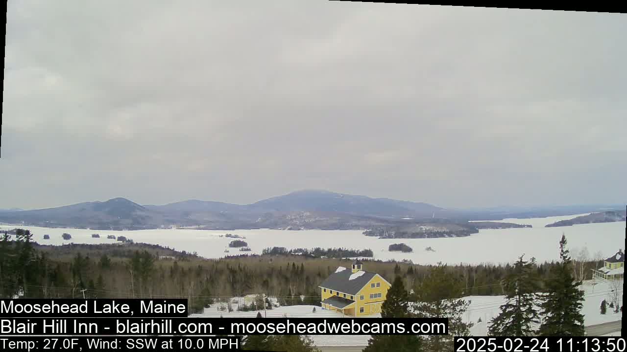 A partially frozen lake is visible from a snowy hilltop, with a yellow building and dark evergreen trees in the foreground, under an overcast sky.