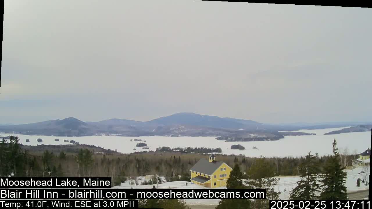 A partially frozen lake is seen from a snowy hillside, with a yellow building and evergreen trees in the foreground, under an overcast sky.