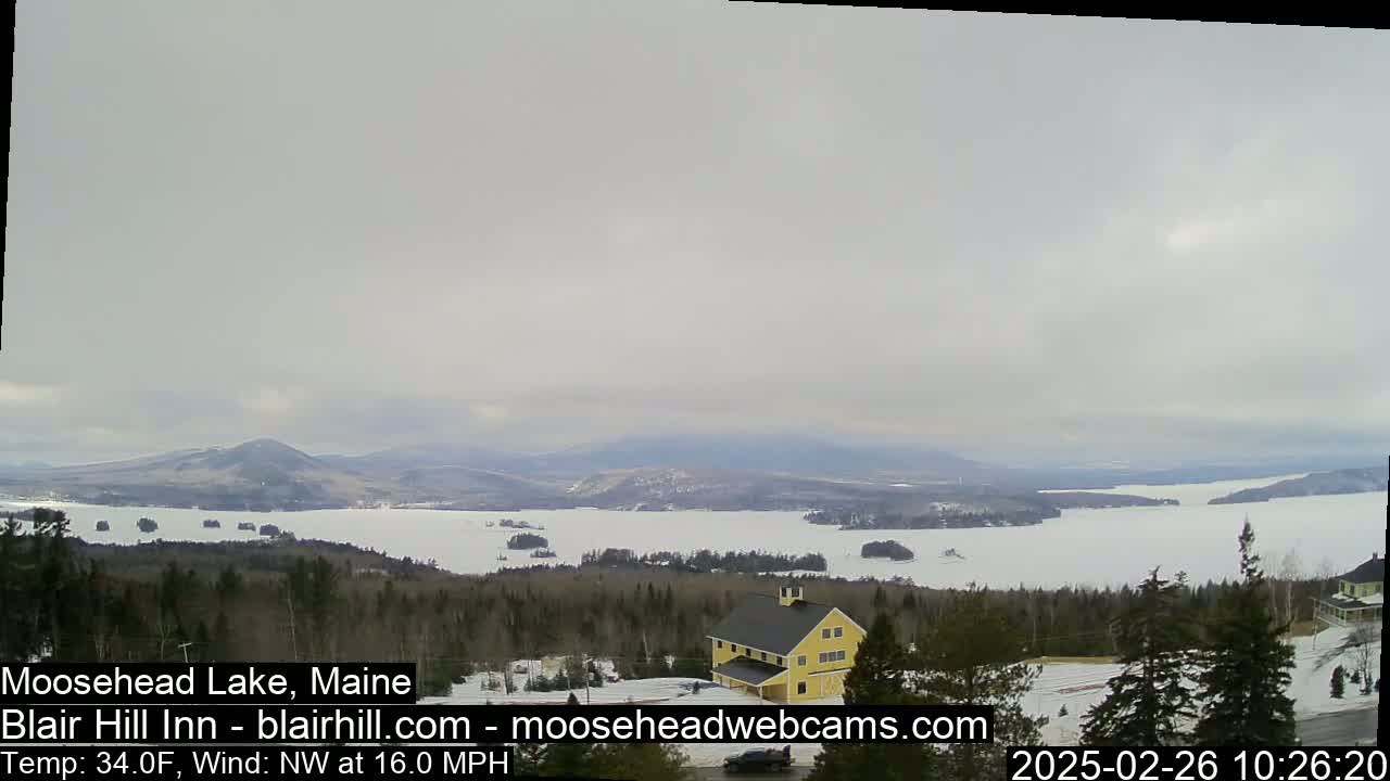 A partially frozen lake stretches to distant mountains under an overcast sky, with a yellow building and snow-covered trees in the foreground.