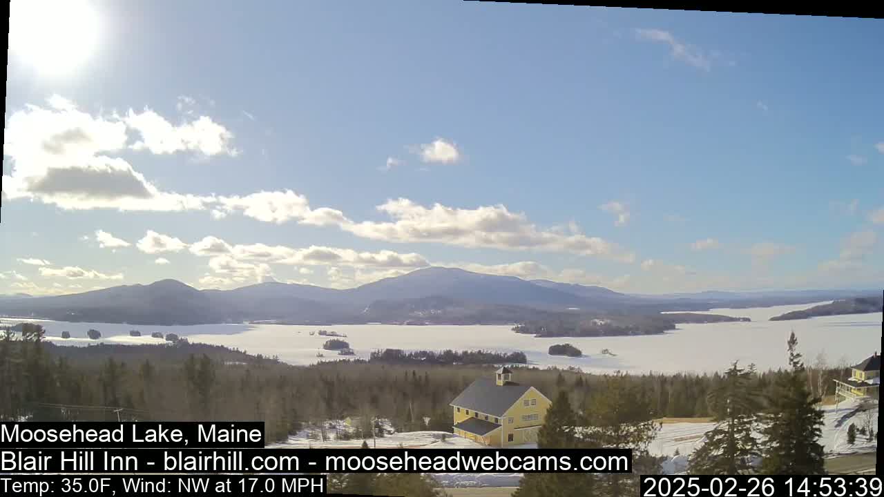 A partly sunny day with a northwest wind of 17 mph shows a snow-covered lake and mountains, viewed from a vantage point overlooking a yellow building.