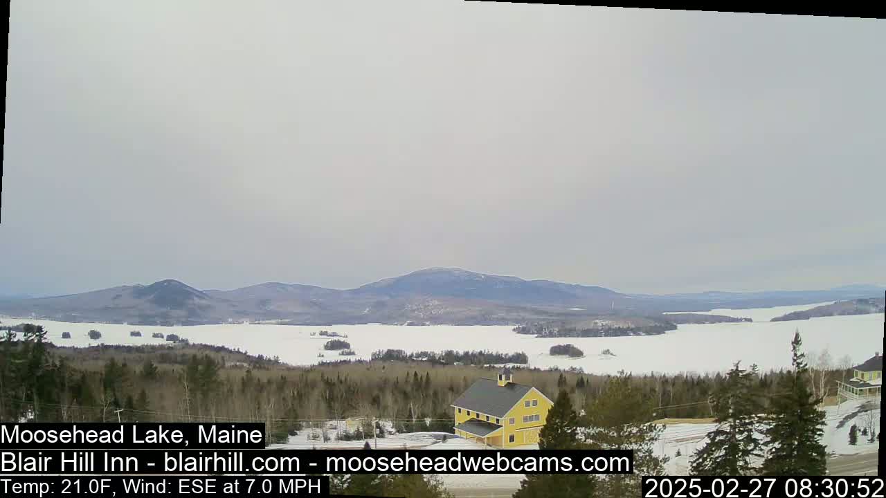 A partially frozen lake stretches to snow-covered mountains under an overcast sky, with a yellow building visible in the foreground.