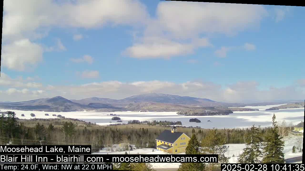 A partly cloudy day reveals a snow-covered lake nestled among forested hills and mountains, with a yellow building visible in the foreground.
