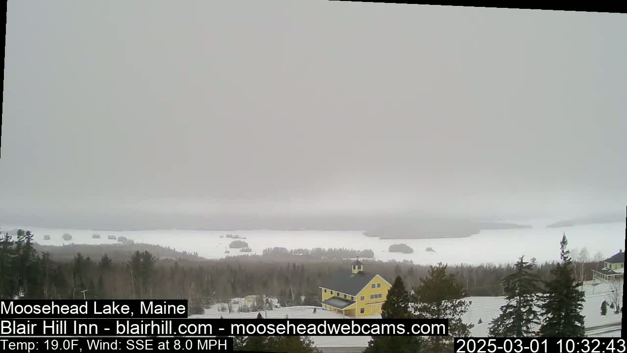 A hazy, snowy landscape shows a partially frozen lake with a yellow building and snow-covered trees in the foreground.