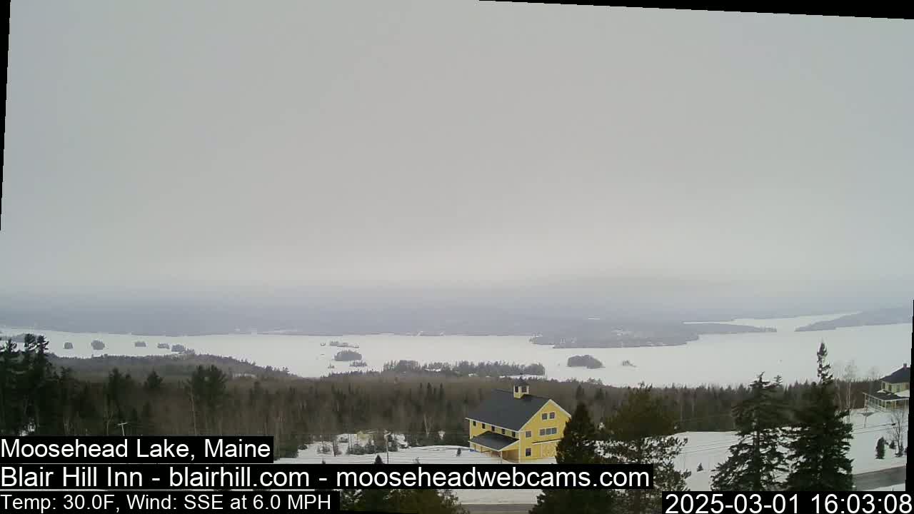 A mostly frozen lake is visible from a snowy hilltop, with a yellow building and evergreen trees in the foreground under an overcast sky.