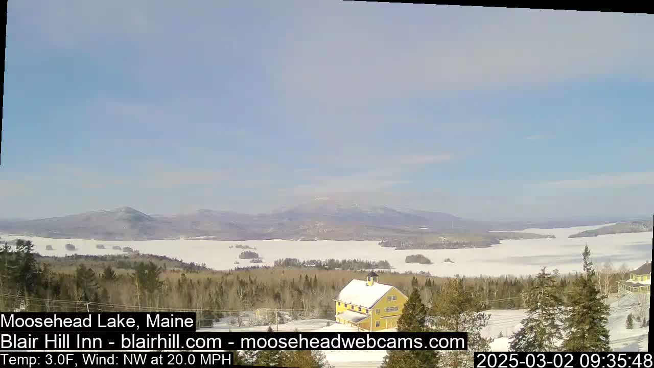 A partly frozen lake stretches to snow-covered mountains under a clear, bright sky, with a yellow building visible in the foreground and a 20 mph northwest wind.