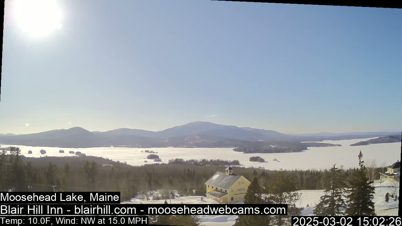 A sunny, cold day with a northwest wind reveals a snow-covered lake and mountains in the distance, with a yellow building situated on a snow-covered hill in the foreground.