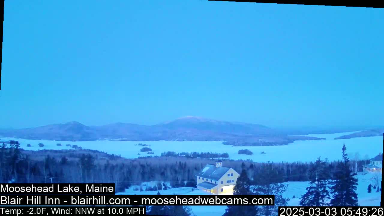 A snow-covered landscape with a frozen lake, distant mountains, and a building, under a clear, twilight sky.