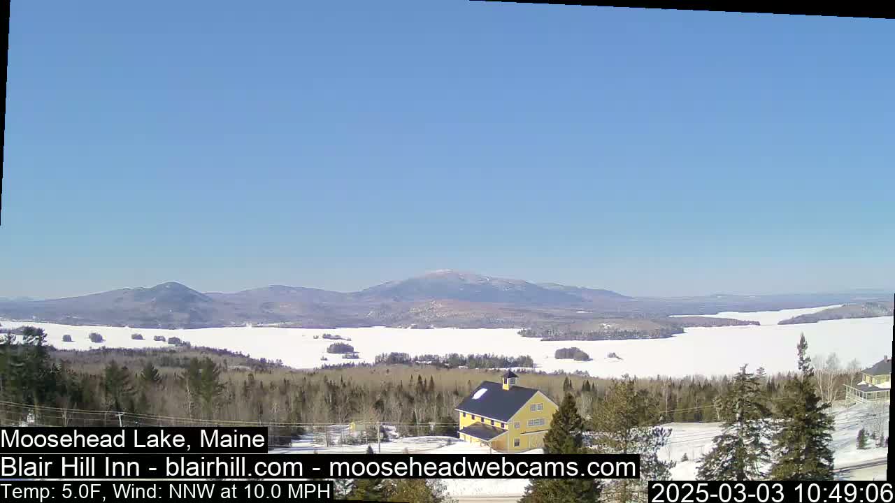 A partially frozen lake and snow-covered landscape are visible under a clear blue sky, with mountains in the background and a yellow building in the foreground.