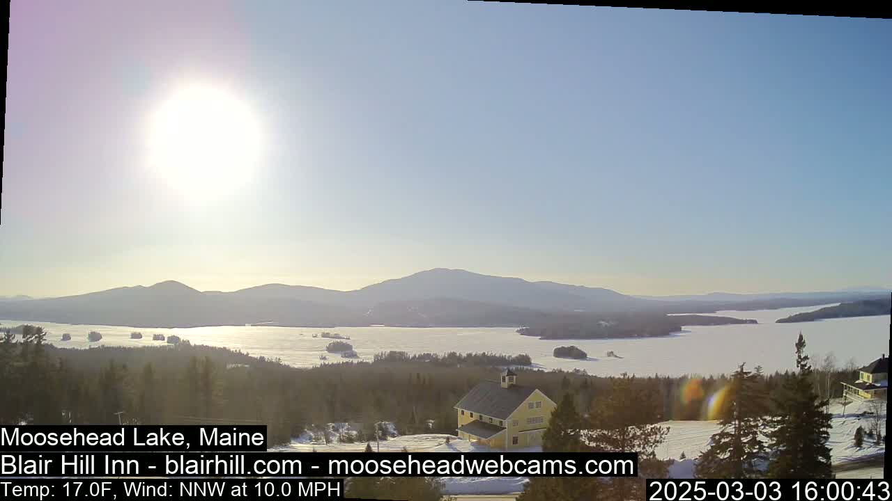 A bright, sunny day reveals a snow-covered lake and distant mountains, with a yellow building nestled in the foreground.