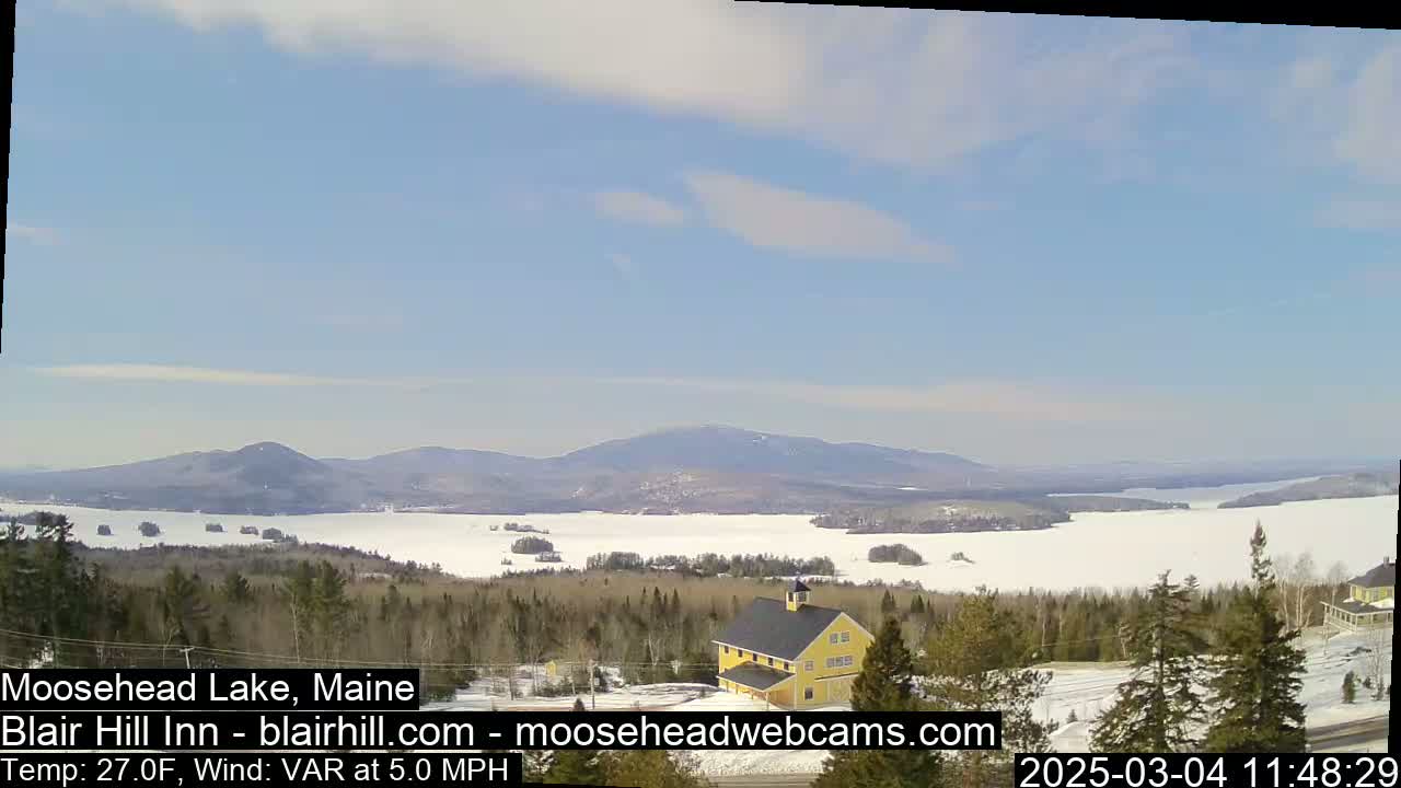 A partly cloudy sky overlooks a snow-covered lake and forest with distant mountains, and a yellow building sits in the foreground.