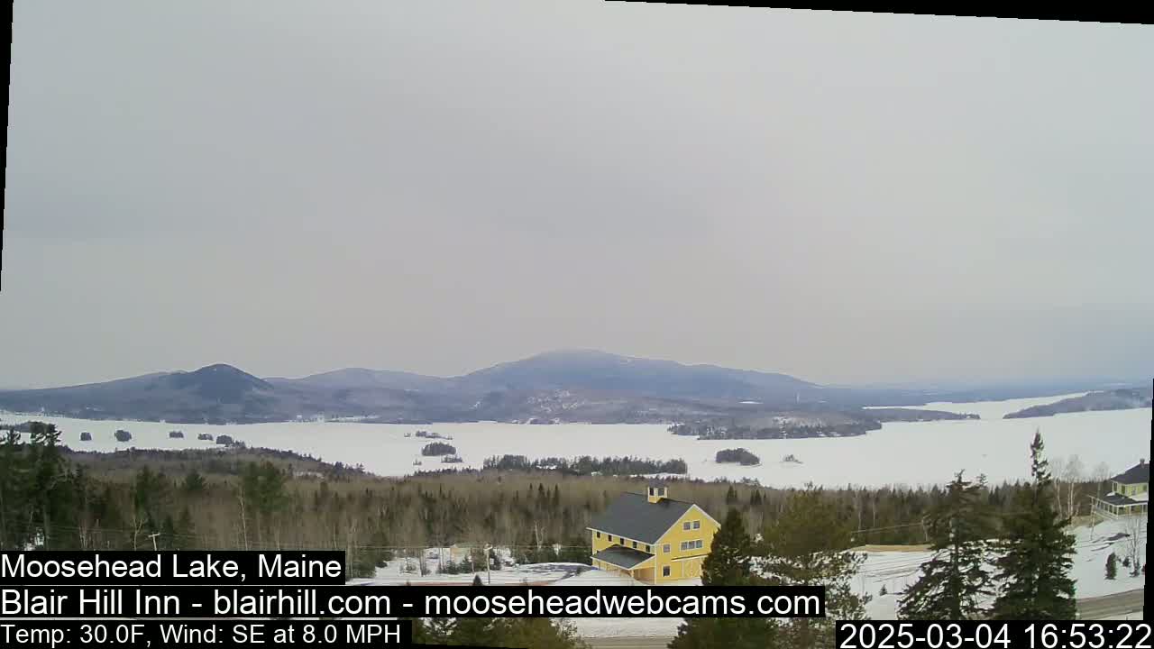 A partly frozen lake stretches to distant mountains under an overcast sky, with a yellow building visible in the foreground.
