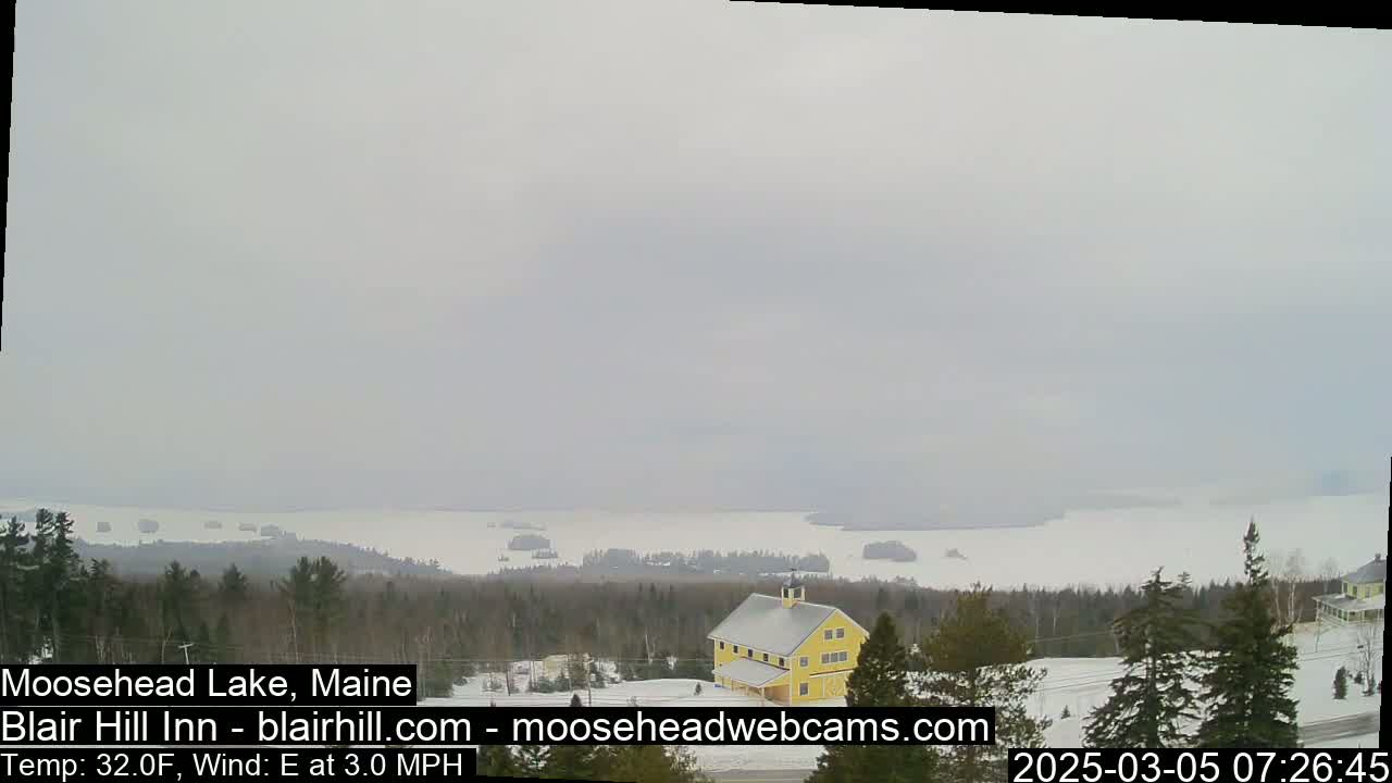 A mostly cloudy, snowy landscape overlooks a large lake with a yellow barn in the foreground.