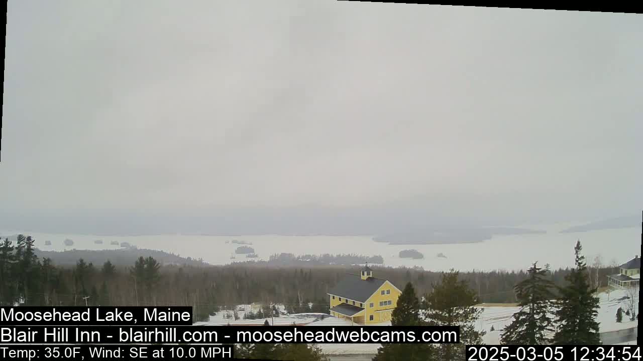 A large lake partially covered in ice is seen from a high vantage point on an overcast day with a 10 mph southeasterly wind, and a yellow building sits in the foreground among some trees and snow.
