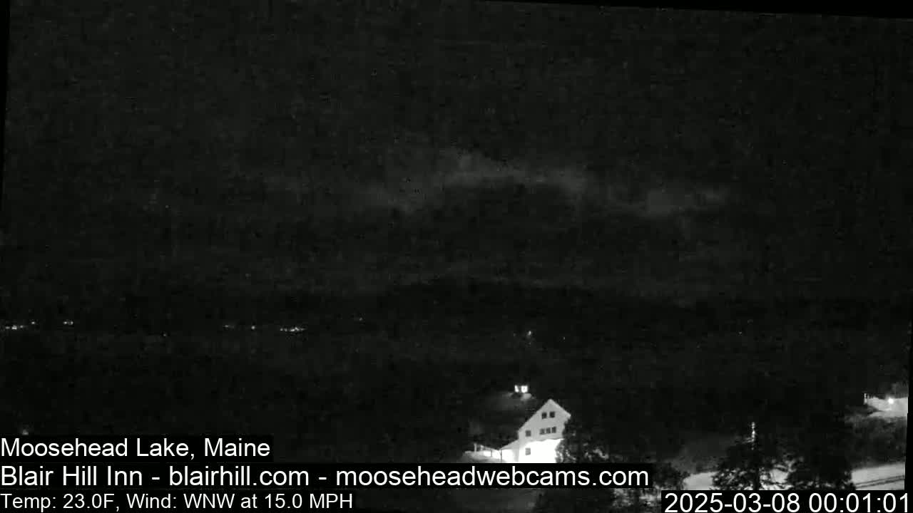 A nighttime, low-light view of a lake and a building under a mostly cloudy sky with a 15 mph wind.