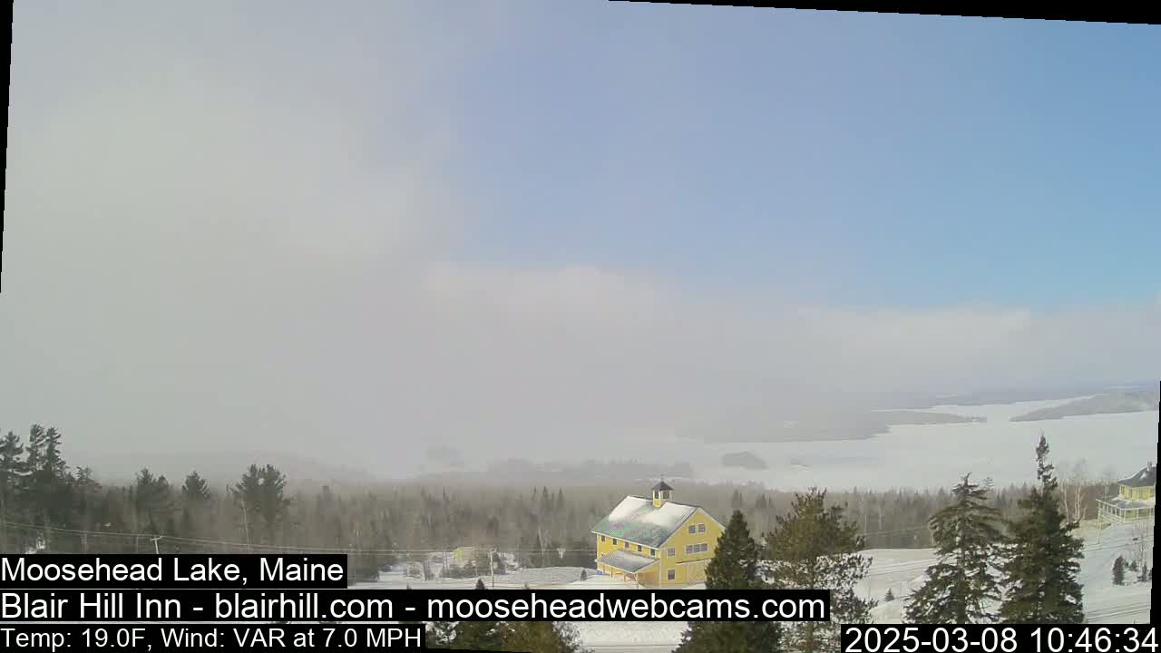 A hazy, partially cloudy day with a 7 mph variable wind shows a snow-covered landscape with a yellow building and a frozen lake in the distance.