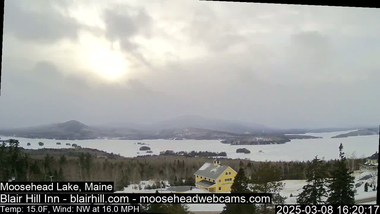 A partially frozen lake is seen from a snowy hilltop, with a yellow building and a partly sunny, somewhat cloudy sky.