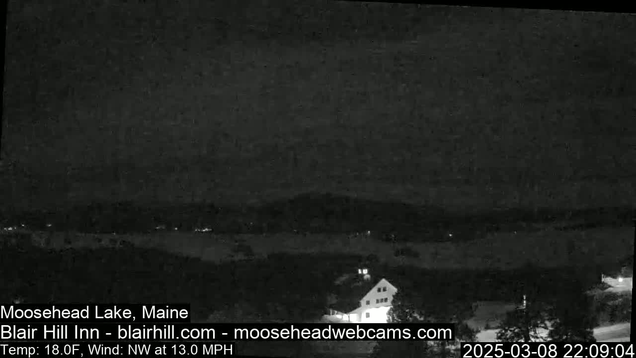 A nighttime, low-light view of a snow-covered landscape with a building and a body of water in the distance, under a dark sky with a northwest wind at 13 mph and a temperature of 18 degrees Fahrenheit.