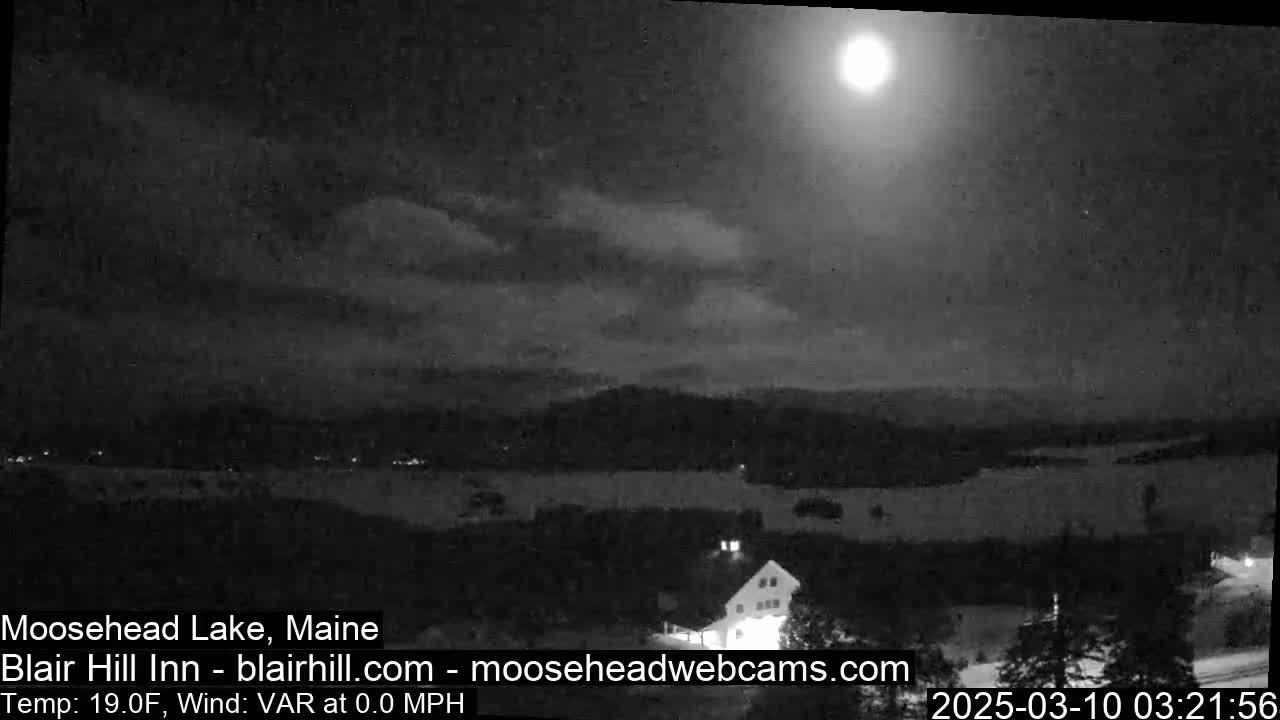 A nighttime view of a snow-covered landscape with a bright moon and some clouds in the sky, overlooking a body of water and a building.