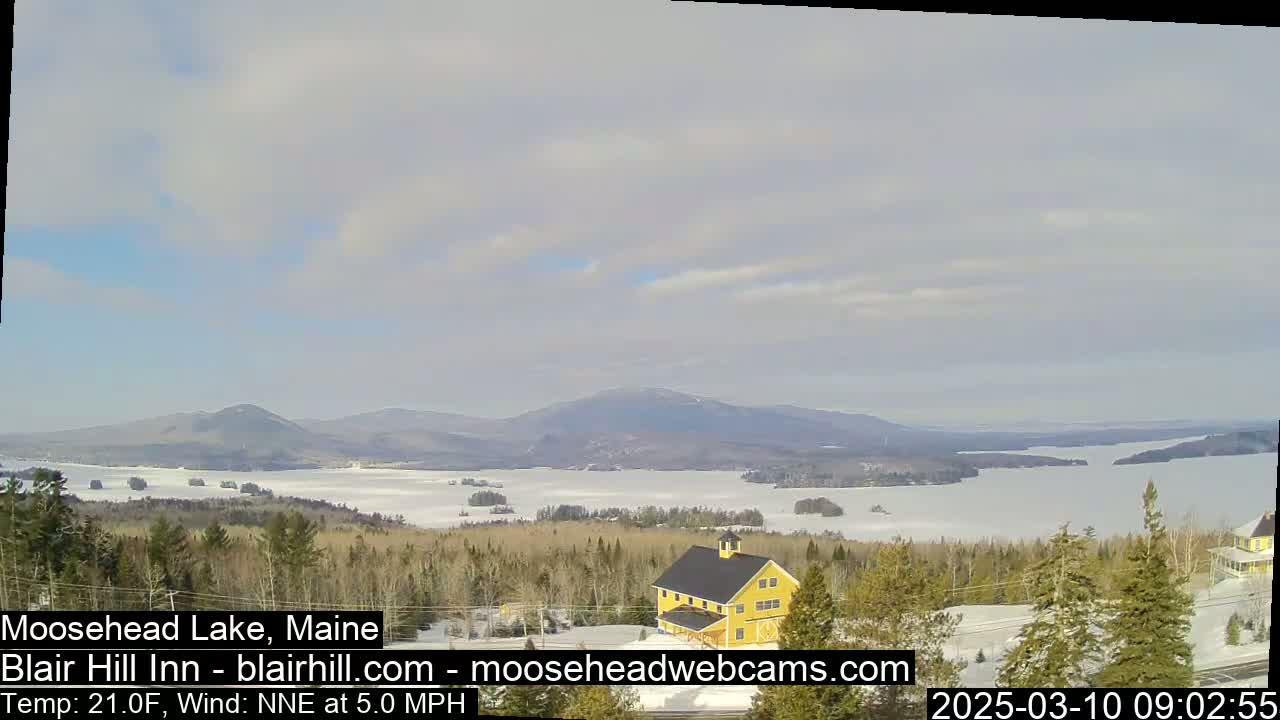 A partly cloudy day reveals a frozen lake and distant mountains, with a yellow building and snow-covered evergreen trees in the foreground.