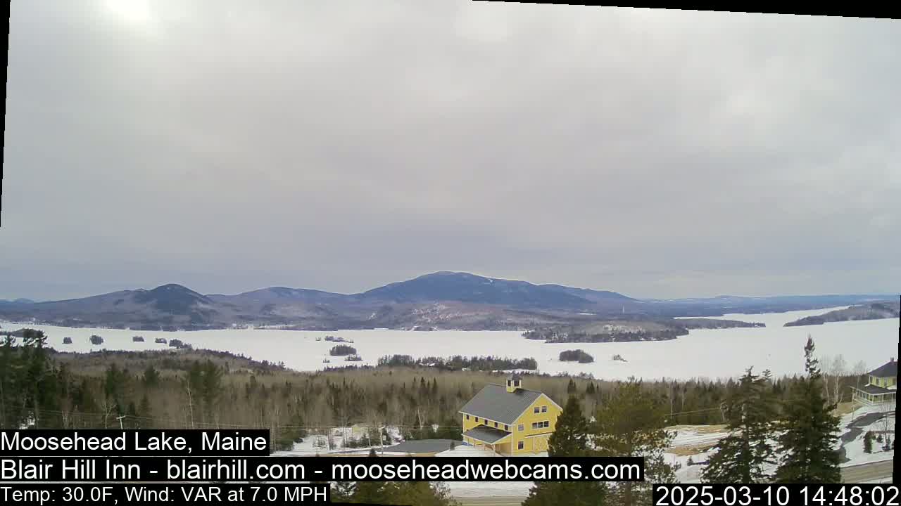 A partly cloudy, cold day shows a partially frozen lake surrounded by snow-covered trees and distant mountains, with a yellow building visible in the foreground.