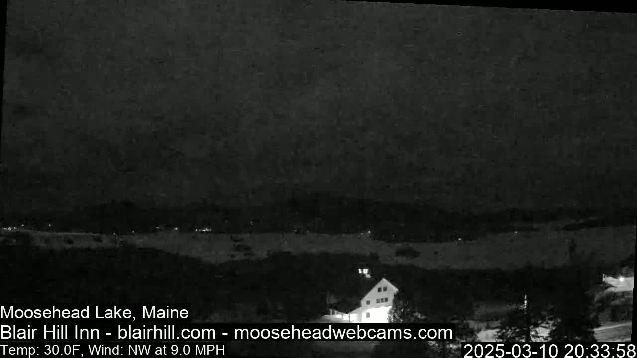 A nighttime view of a lake and surrounding land, with a building visible in the bottom right corner, under a dark sky with a northwest wind at 9 mph and a temperature of 30°F.