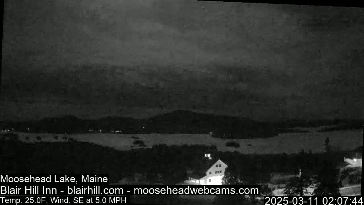 A nighttime, low-light view of a snow-covered landscape with a lake and distant mountains under a cloudy sky, with a light wind from the southeast.