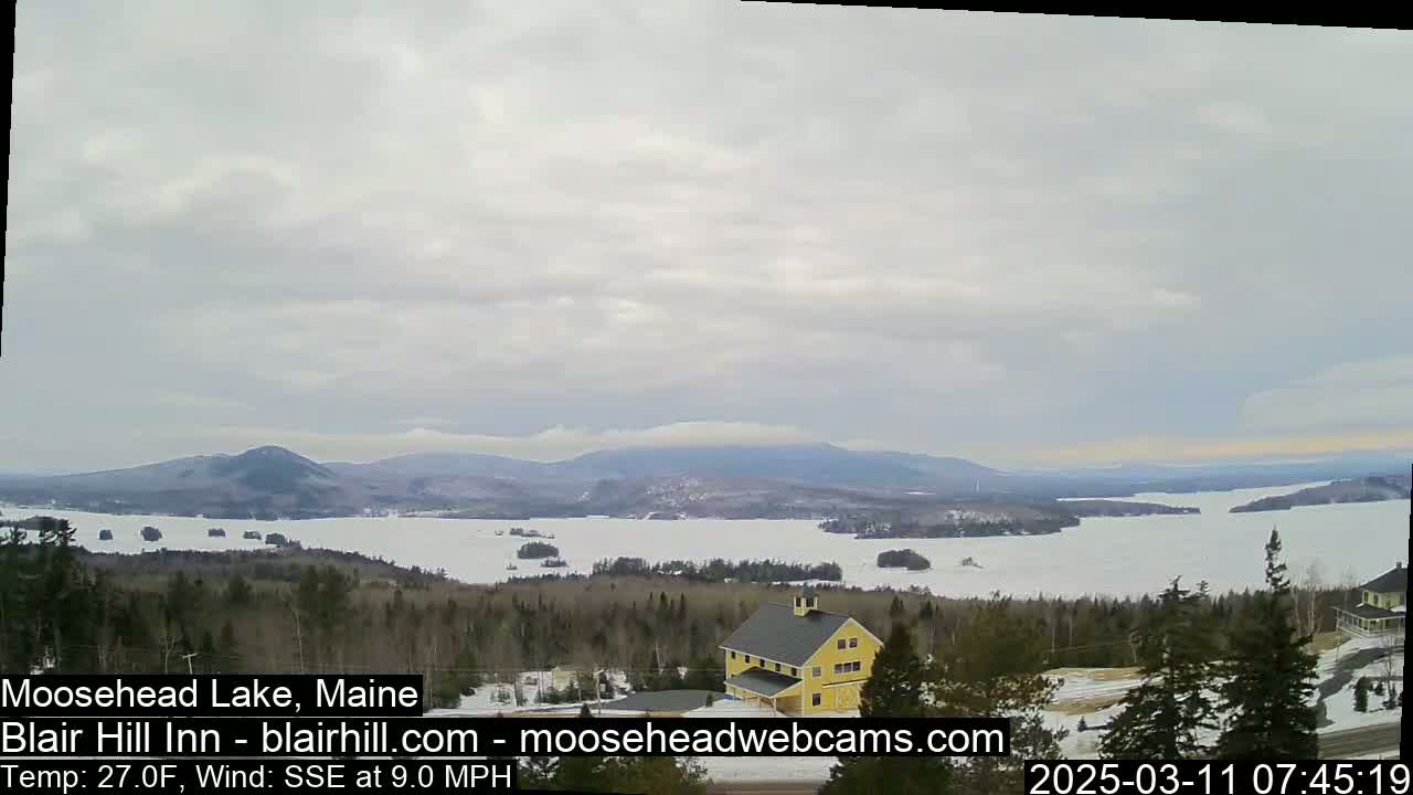 A partly frozen lake stretches to distant, snow-dusted mountains under a cloudy sky, with a yellow building visible in the foreground.