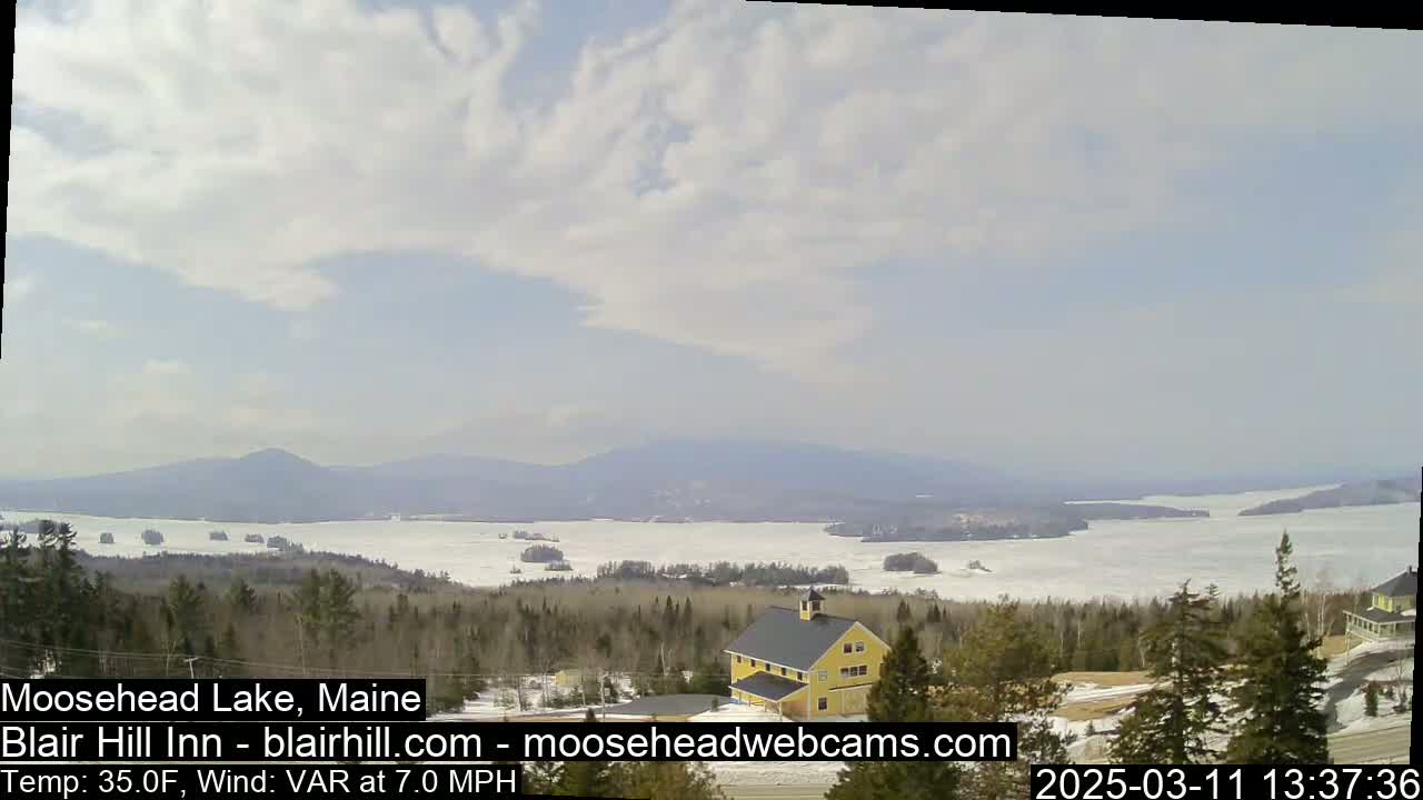 A partly cloudy day overlooks a mostly frozen lake with distant mountains, a yellow building, and evergreen trees in the foreground.