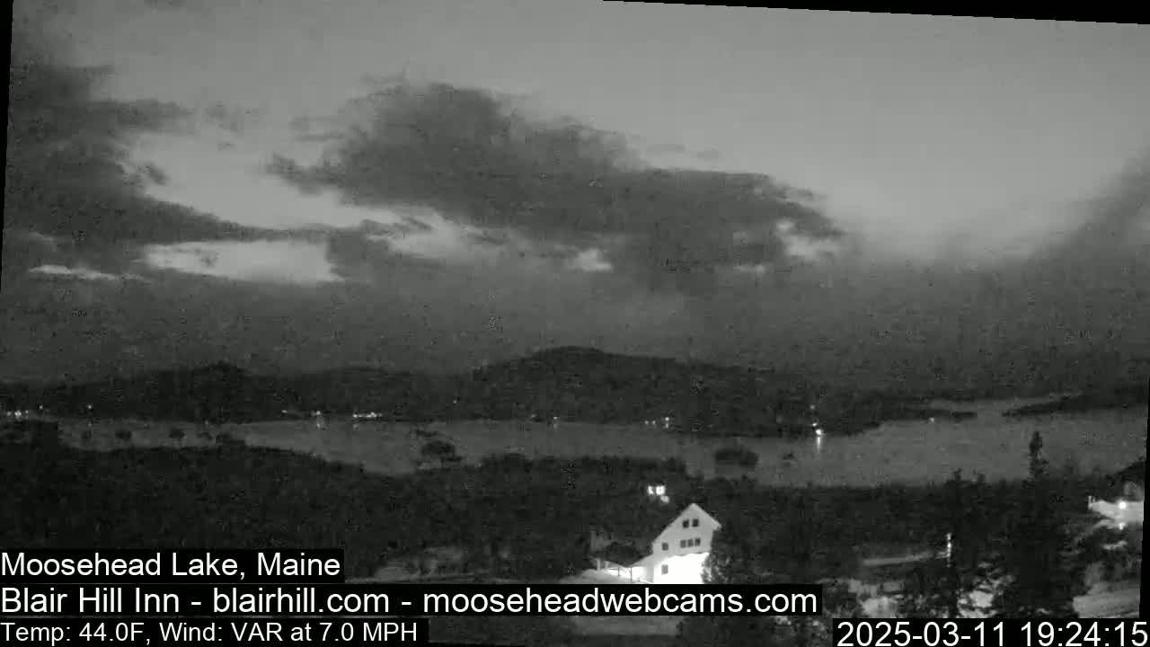 A nighttime, overcast view of a lake and distant mountains under a cloudy sky with a 7.0 mph variable wind and a temperature of 44°F.