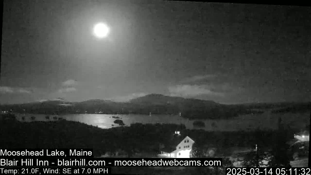 A bright moon illuminates a calm lake at night, with dark mountains in the background under a partly cloudy sky.