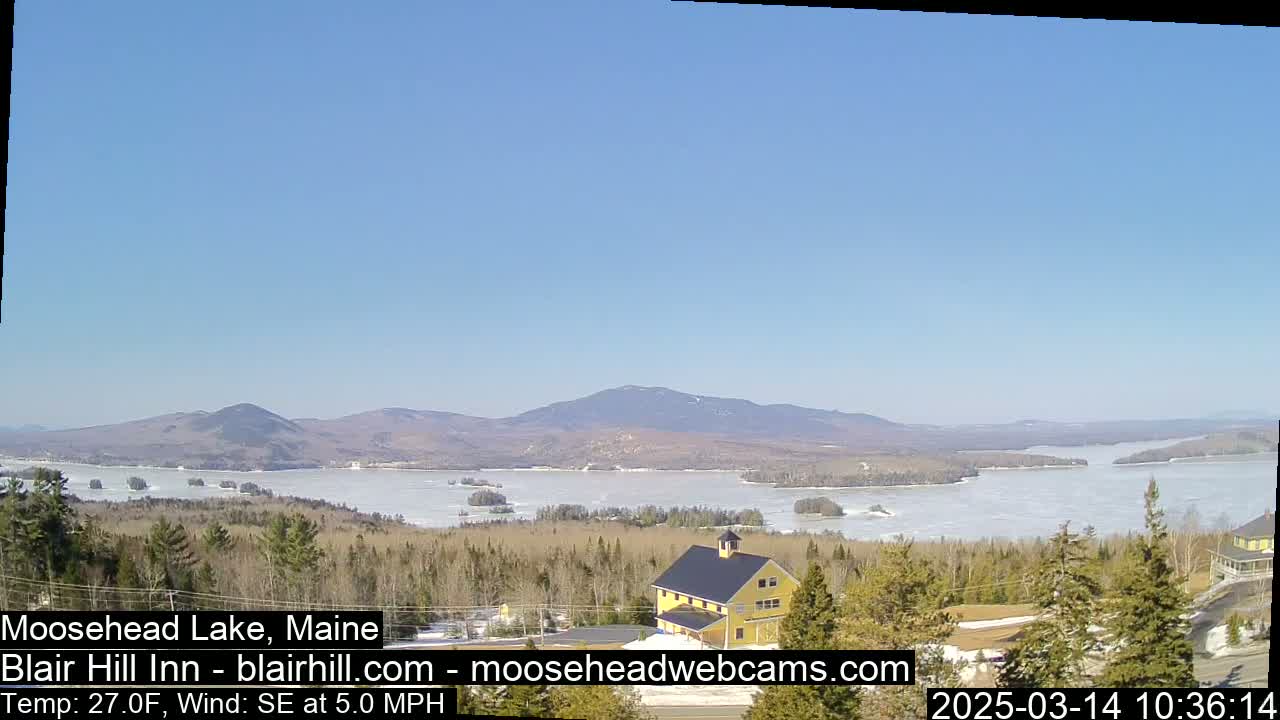 A partly frozen lake stretches to the base of distant mountains under a clear blue sky, with a yellow building and evergreen trees in the foreground.