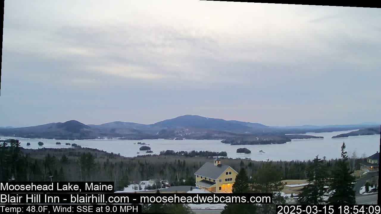 A partly cloudy sky overlooks a large lake partially frozen, with mountains in the distance and a yellow building nestled in the trees in the foreground.