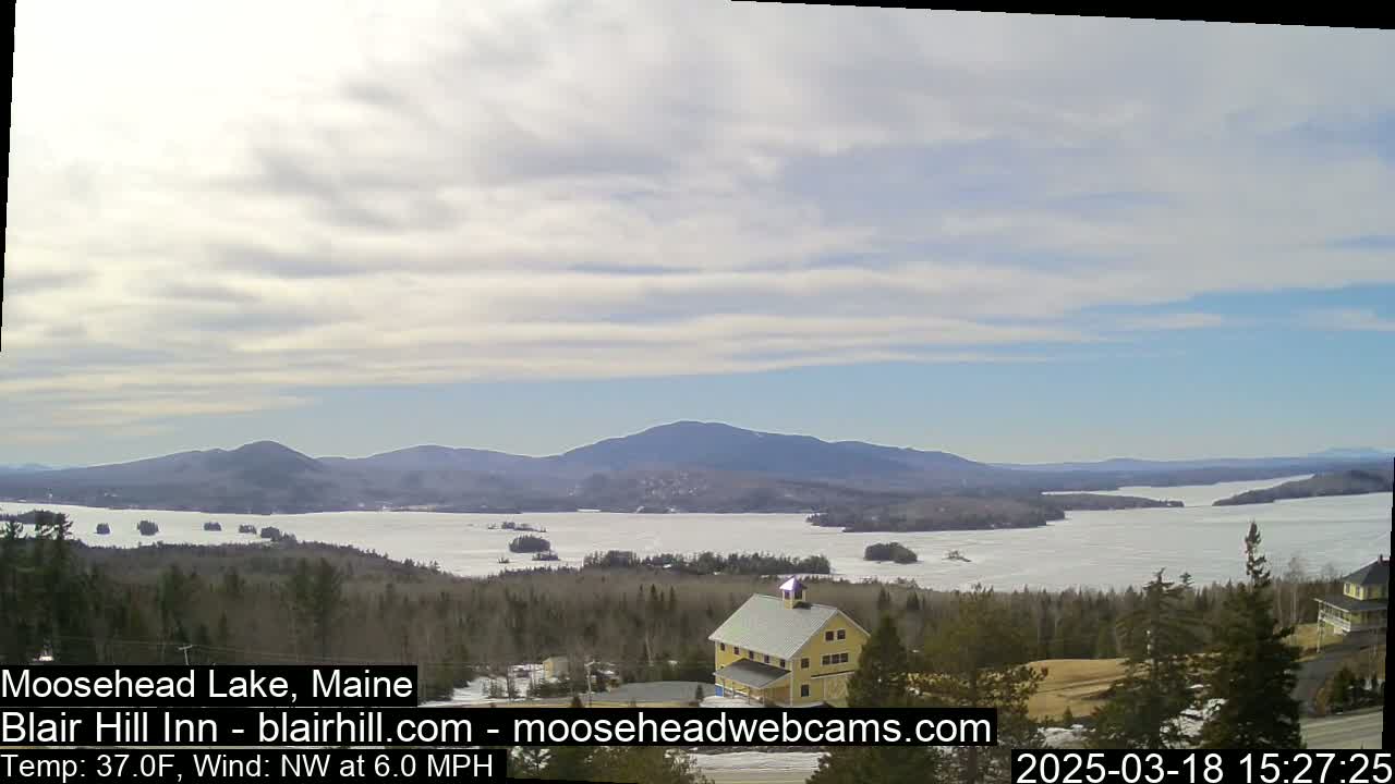 A partly cloudy day reveals a frozen lake surrounded by snow-covered hills and evergreens, with a yellow building visible in the foreground.