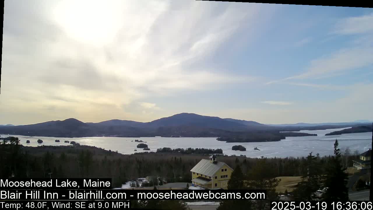 A partly cloudy day reveals a view of a large lake partially frozen, surrounded by dark green trees and distant mountains, with a yellow building nestled in the foreground.