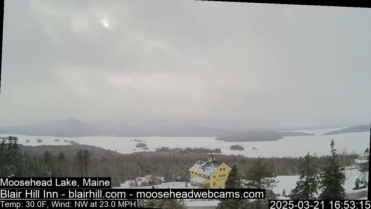 A snow-covered landscape shows a partially frozen lake under an overcast sky, with a yellow building nestled among evergreens in the foreground.