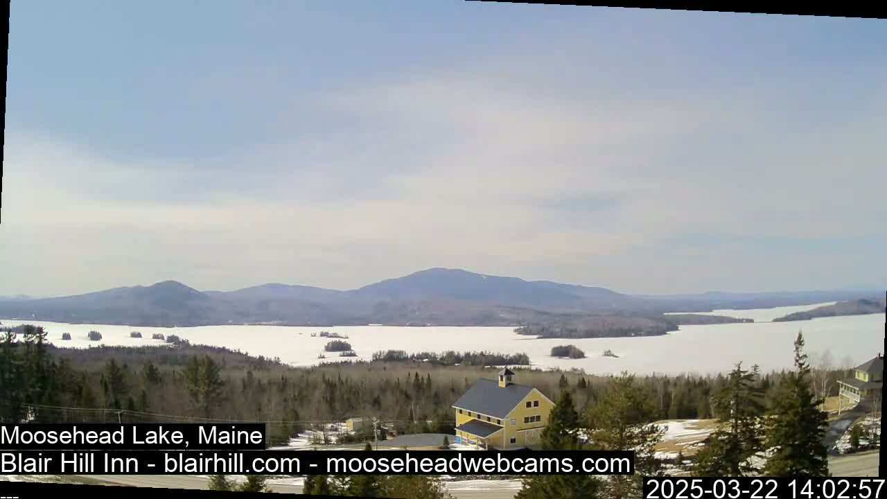 A partly sunny day reveals a snow-covered lake stretching to distant mountains, with a yellow building visible in the foreground.