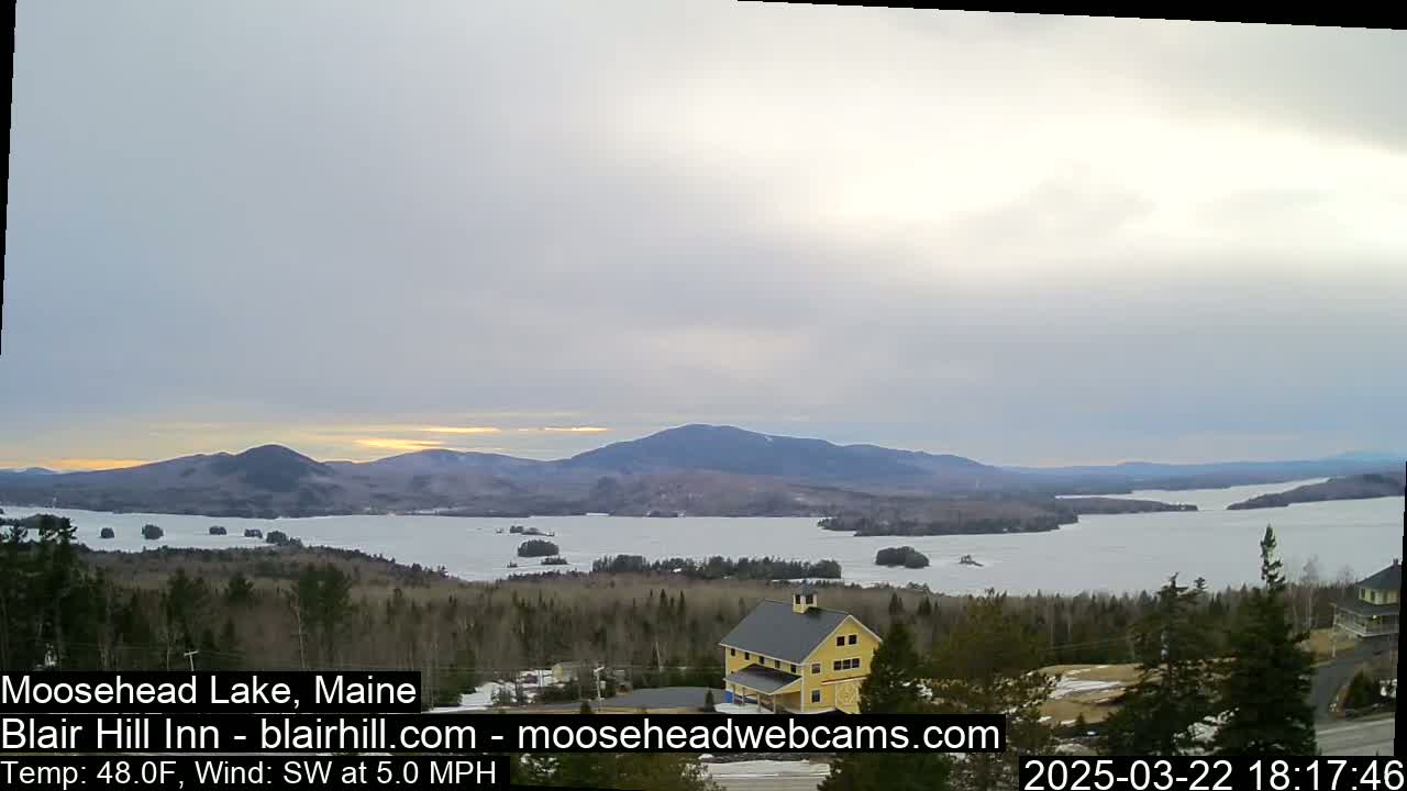 A partly cloudy sunset over a partially frozen lake and mountains, viewed from a wooded hillside with a yellow building in the foreground.