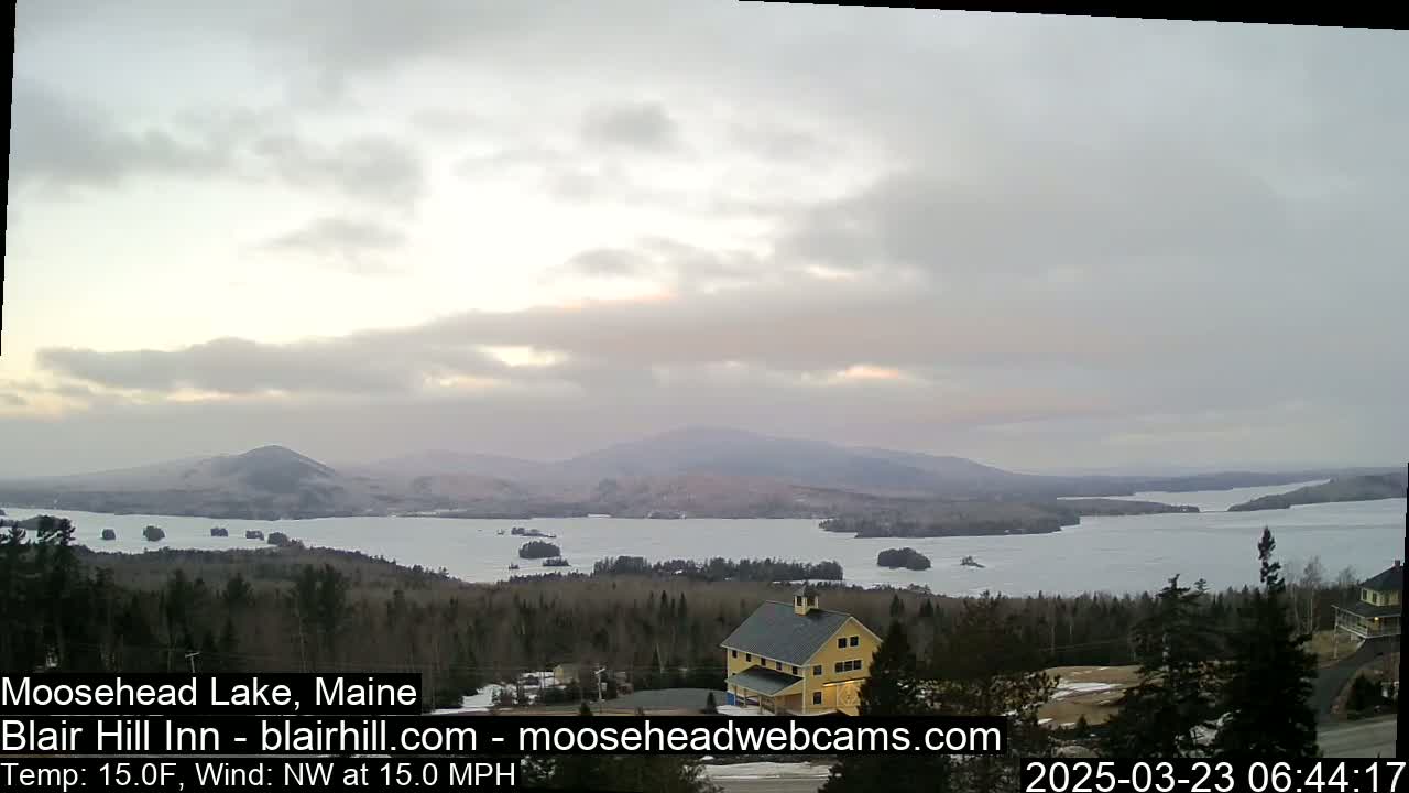 A partially frozen lake is surrounded by snow-covered hills and evergreen trees under an overcast sky.