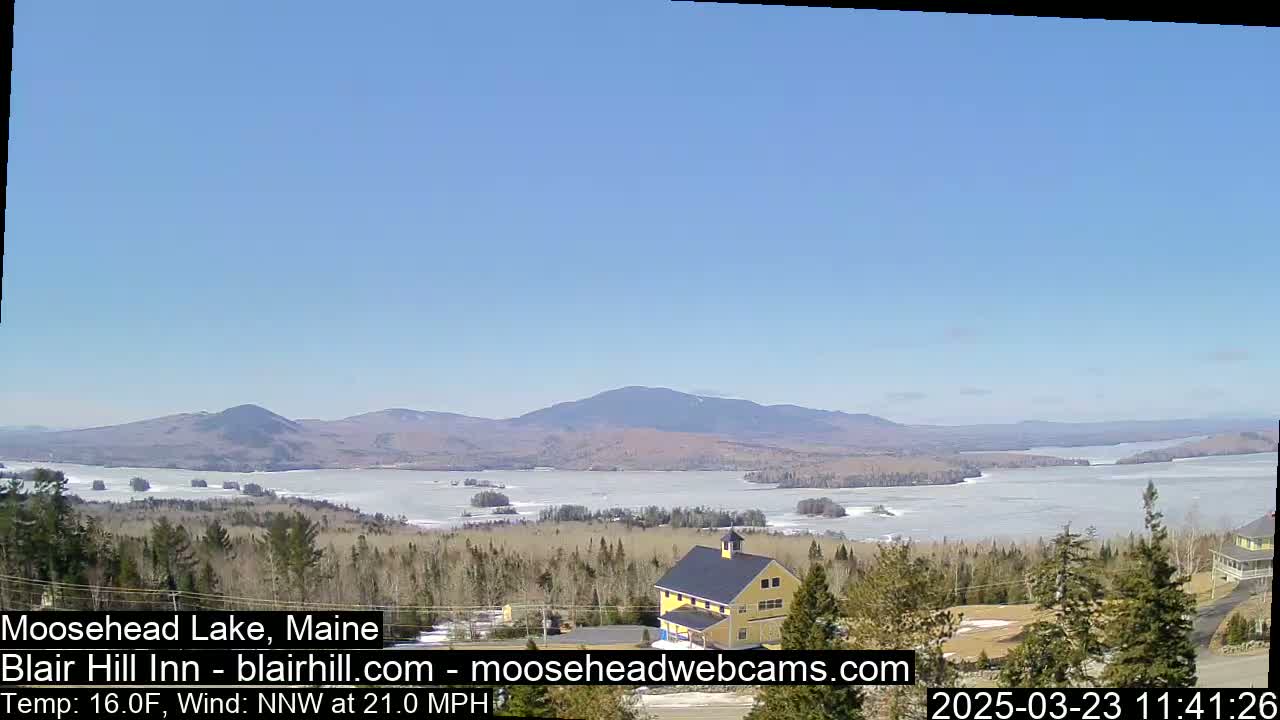 A partly frozen lake stretches to distant mountains under a clear blue sky, with a yellow building visible in the foreground and a brisk north-northwest wind.