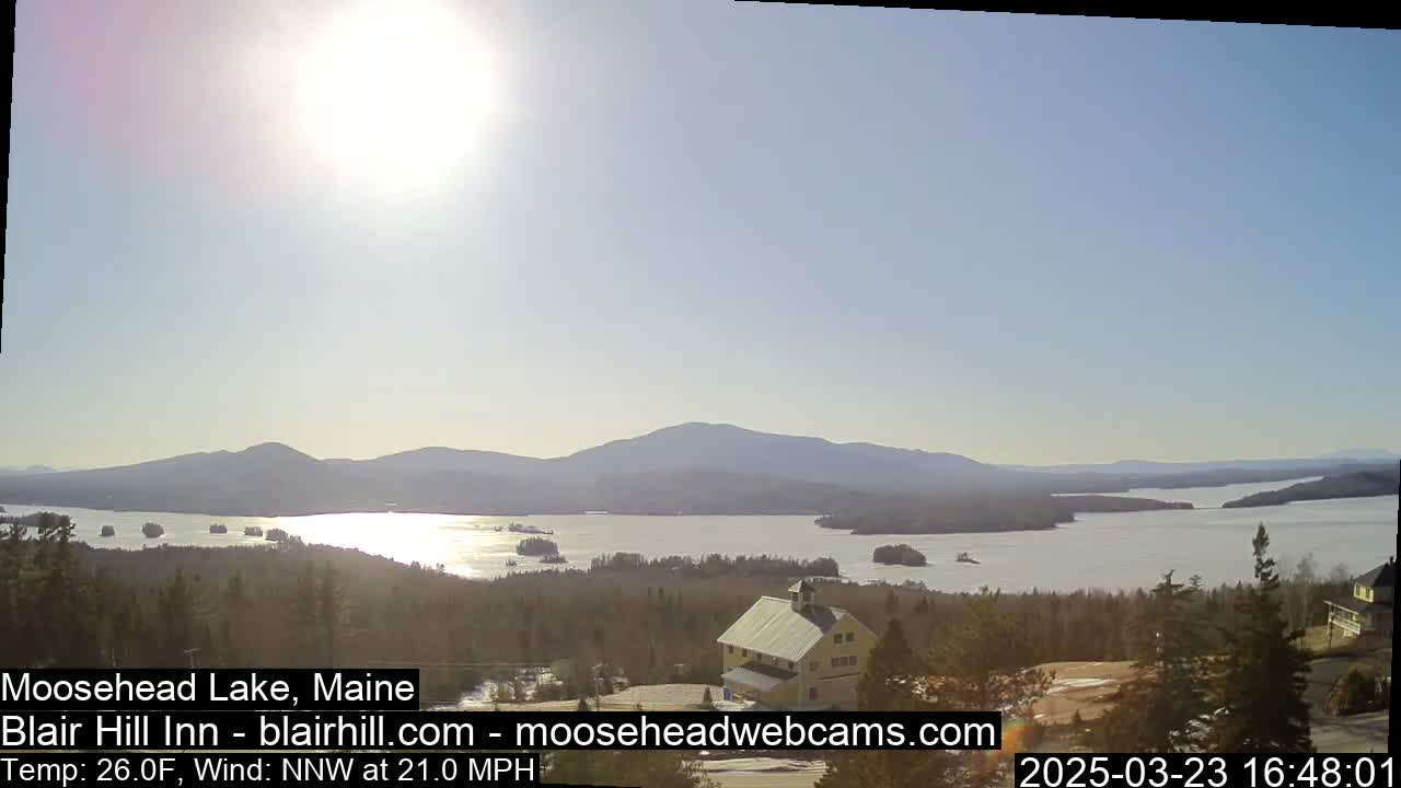 A sunny, bright day with a northwest wind shows a large lake surrounded by mountains and evergreen trees, with a small building visible in the foreground.