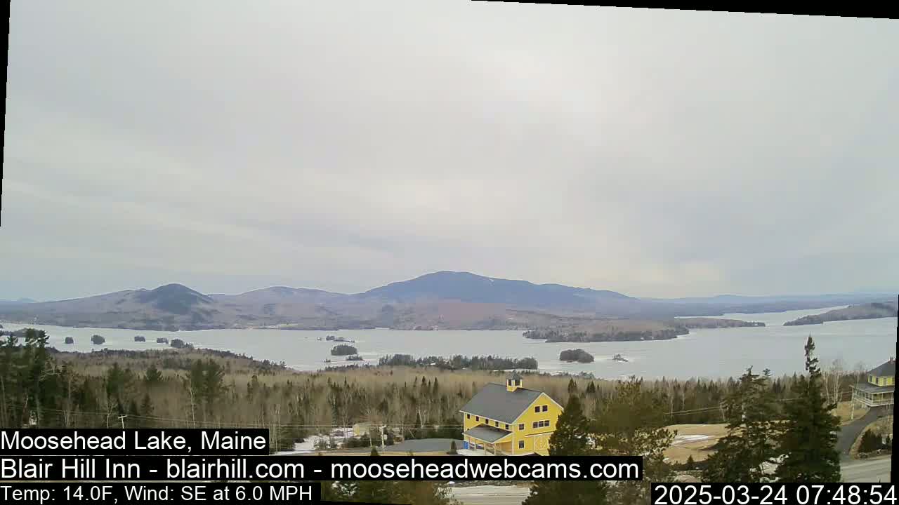 A partly frozen lake is seen from a high vantage point on a cloudy day, with a yellow building and distant mountains in view.