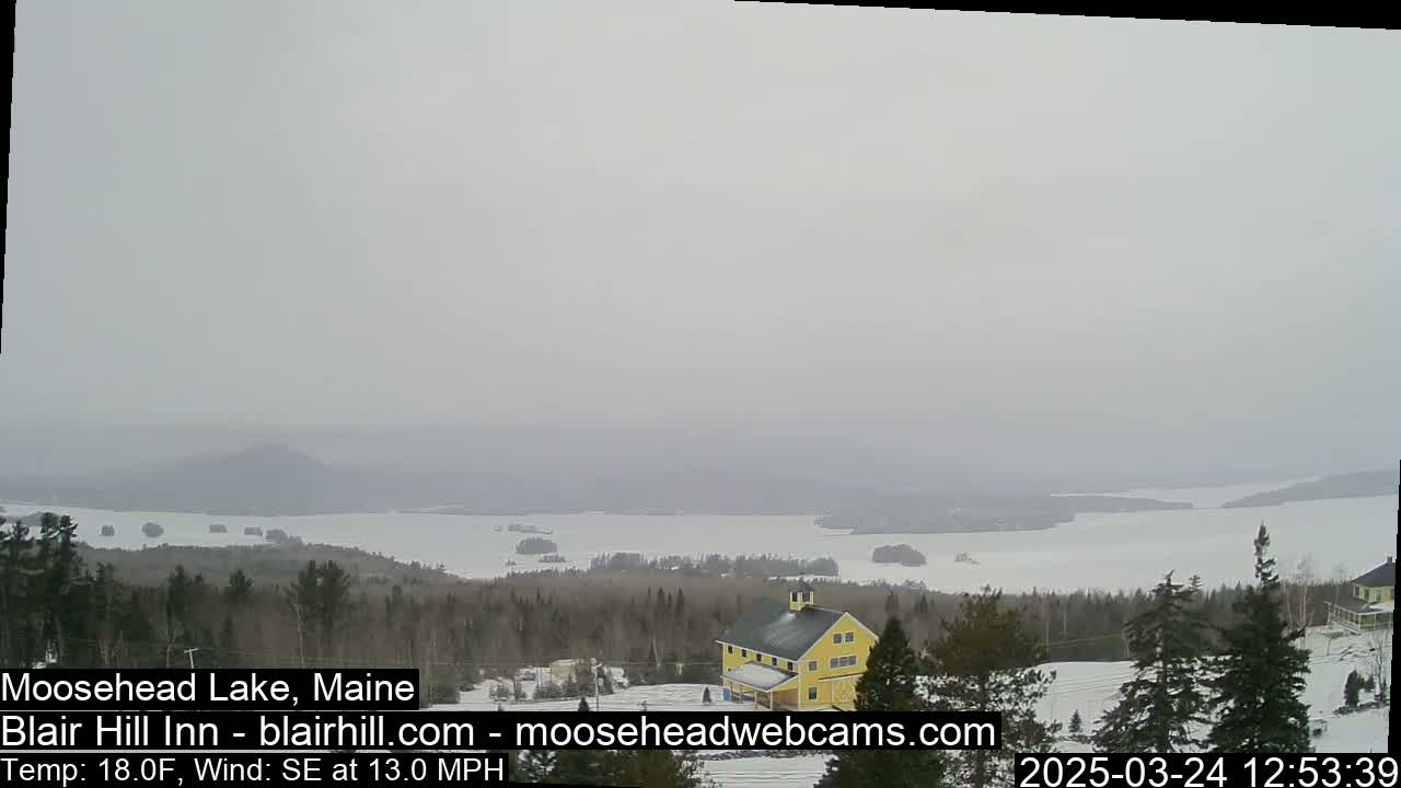 A hazy, snowy landscape shows a partially frozen lake and a yellow building nestled among evergreens.
